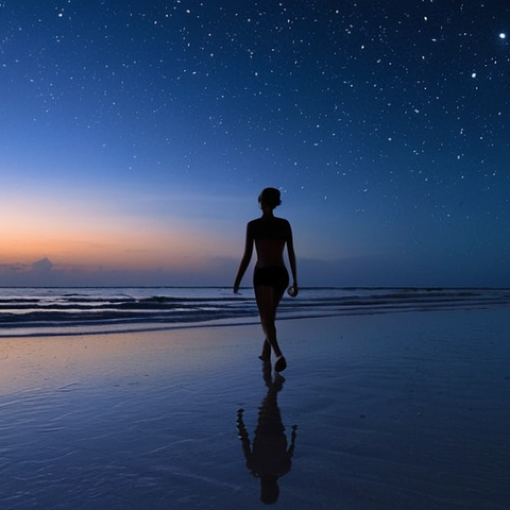 **Holbox, Mexico:** A serene beach scene at dusk with bioluminescent microorganisms illuminating the shoreline, resembling a starry sky reflecting on the sea. A silhouette of a person walking along the water's edge.